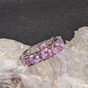 May include: A silver ring with a continuous row of square-cut, light pink gemstones. The ring is set on a white, crystalline surface against a dark background. The gemstones are evenly spaced and reflect light.