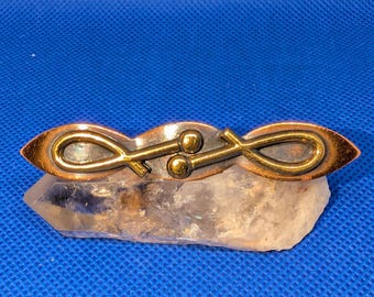 May include: A horizontal metal brooch featuring a polished brass-toned finish with two interconnected loop shapes and two small spherical accents in the center, resting atop a translucent, jagged, clear quartz crystal formation.