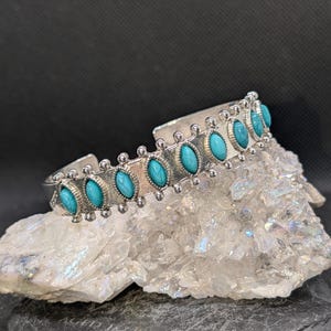 May include: A white ceramic mug with a black handle and the text "But First, Coffee" in a bold, black font. The mug is set against a blurred background, suggesting a cozy setting.