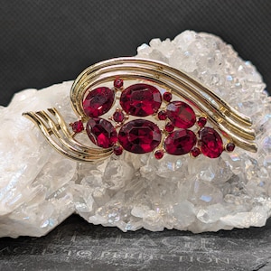 May include: A gold-tone metal brooch featuring a curved, sweeping design accented with multiple oval-shaped, deep red faceted stones. The brooch is arranged in a cluster of larger and smaller red gems set within polished gold-colored metal ribbons.