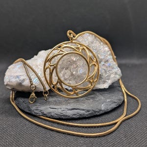 May include: A gold-toned necklace featuring a circular pendant with an intricate, openwork swirling pattern. The pendant is suspended from a thin, flexible snake chain with a lobster claw clasp, resting against a rough, translucent white crystal.