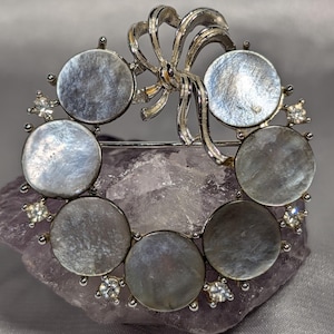 May include: A circular brooch featuring seven iridescent, round mother-of-pearl discs arranged in a wreath pattern. The silver-toned metal frame includes a decorative bow at the top and small, clear, faceted rhinestones set between the discs.