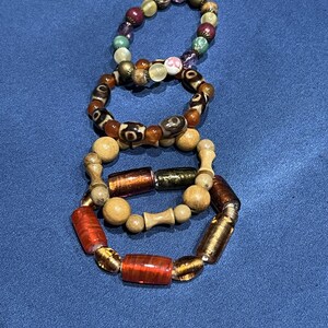 May include: Three beaded bracelets with a variety of beads in different colors and shapes. The top bracelet has purple, green, and gold beads. The middle bracelet has brown and tan beads. The bottom bracelet has red and gold beads.