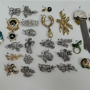 May include: A collection of vintage brooches in various designs, including floral, bow, and abstract shapes. The brooches feature rhinestones, gold, and enamel accents. Some have dangling elements, and others are shaped like animals.