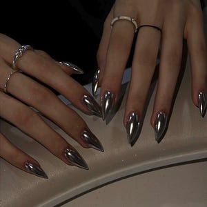 Puede incluir: Primer plano de manos con uñas largas plateadas cromadas en forma de estilete. Las uñas tienen un acabado metálico reflectante. Las manos también tienen anillos plateados en los dedos. El fondo es oscuro.