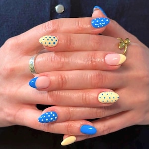 Könnte beinhalten: Nahaufnahme einer Hand mit verschiedenen Nageldesigns. Einige Nägel sind blau mit weißen Punkten, andere sind einfarbig blau oder gelb. Ein Nagel hat eine rosa Basis mit einer gelben Spitze. Ein goldener Ring ist sichtbar.