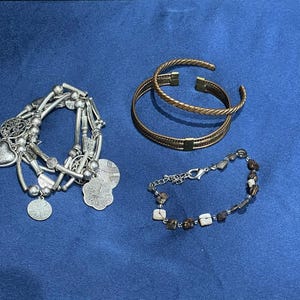 Puede incluir: Cuatro pulseras sobre una superficie azul. Una es plateada con abalorios, incluyendo corazones y colgantes redondos. Dos son doradas, una con un diseño trenzado. La cuarta pulsera está hecha con cuentas cuadradas marrones y blancas.