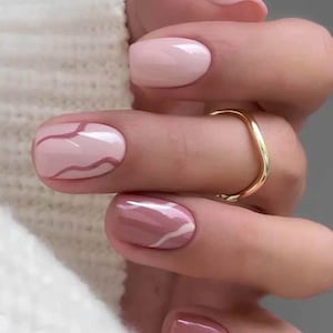 Può includere: Primo piano di una mano con un anello d'oro e una manicure con varie tonalità di rosa. Alcune unghie sono rosa pieno, mentre altre hanno un design marmorizzato con accenti bianchi. Le unghie sono di forma ovale.