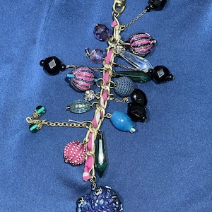 Op de afbeelding: Een decoratieve sleutelhanger met een goudkleurige sluiting en ketting. De sleutelhanger heeft een verscheidenheid aan kleurrijke kralen, waaronder roze, paars, zwart en blauw, samen met een roze lint en een grote, getextureerde blauwe hartvormige bedel.