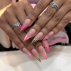 Puede incluir: Uñas acrílicas largas y puntiagudas con un diseño de manicura francesa. Las puntas están decoradas con lunares rosas y amarillos. Se ven dos anillos plateados en los dedos.