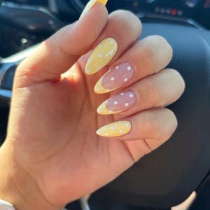 May include: A hand with long, almond-shaped nails. The nails feature a design with a yellow base and white polka dots, with a French tip in yellow. Some nails have a pink base with white polka dots.