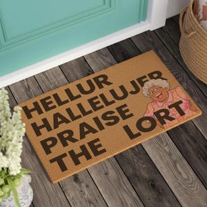 May include: A rectangular tan coir doormat featuring the black text HELLUR HALLELUJER PRAISE THE LORT and a colorful illustration of an elderly person with gray hair wearing a pink floral shirt and glasses, positioned on a dark wooden floor.