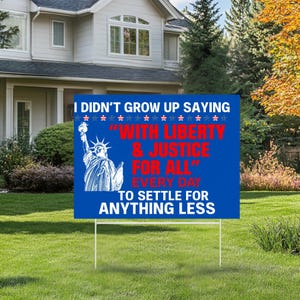 May include: A blue yard sign with white text that reads "I DIDN'T GROW UP SAYING" at the top. Below, in red, it says "WITH LIBERTY & JUSTICE FOR ALL" and "EVERY DAY TO SETTLE FOR ANYTHING LESS". A graphic of the Statue of Liberty is on the left.