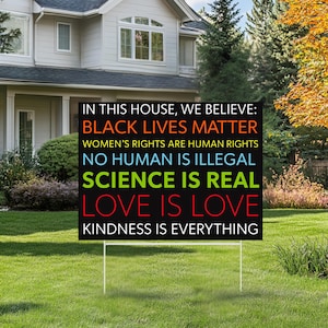 May include: A black yard sign with white stakes, displaying colorful text: "IN THIS HOUSE, WE BELIEVE: BLACK LIVES MATTER, WOMEN'S RIGHTS ARE HUMAN RIGHTS, NO HUMAN IS ILLEGAL, SCIENCE IS REAL, LOVE IS LOVE, KINDNESS IS EVERYTHING."