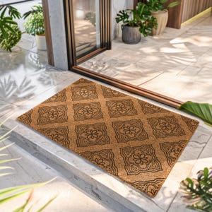 May include: A rectangular natural tan coir doormat featuring a repeating black geometric pattern of square tiles with central floral motifs. The mat has a coarse, fibrous texture and is positioned on a light-colored tiled floor surface.