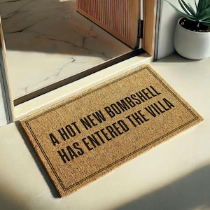 May include: A rectangular tan coir doormat with a black border and the black text A HOT NEW BOMBSHELL HAS ENTERED THE VILLA printed in the center. The mat is positioned on a light-colored floor in front of a doorway with a wooden frame.