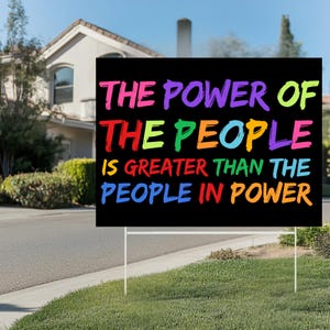 May include: A black yard sign with the text "THE POWER OF THE PEOPLE IS GREATER THAN THE PEOPLE IN POWER" in various bright colors. The sign is displayed on a green lawn in front of a house.