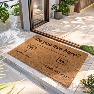 May include: A rectangular brown coir doormat featuring a black printed flowchart graphic. The text reads: Do you live here? with two branches: Yes, leading to welcome home, and No, leading to come back with pizza. The mat has a coarse texture.