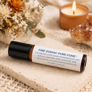 May include: A roll-on essential oil bottle with a black cap and a label that reads "FIRE ZODIAC PURE-FUME". The bottle is on a stone surface, with a lit candle and dried flowers in the background. The bottle contains 10 mL of oil.