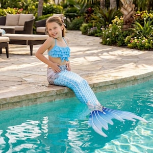 Può includere: Un costume da bagno a due pezzi azzurro composto da un top bikini con volant e una gonna a coda di sirena con un motivo a squame stampato che sfuma dall'azzurro in vita a un blu più scuro sulla pinna ampia e svasata sul fondo.