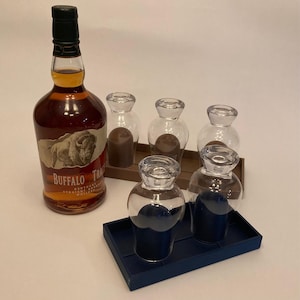 May include: A bottle of Buffalo Trace Kentucky Straight Bourbon Whiskey next to several clear glass decanters and a dark blue tray. The decanters have a dark blue or brown liquid inside. The bottle has a black cap and a label with a bison illustration.