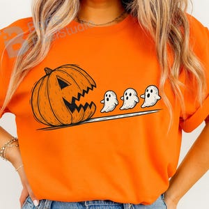 Puede incluir: Camiseta naranja brillante con un diseño de Halloween. El gráfico muestra una calabaza tallada con una boca dentada y tres figuras de fantasmas blancos. La obra de arte en blanco y negro contrasta con la camiseta naranja.