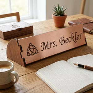 Puede incluir: Una placa de escritorio de madera con una parte frontal de color cobre, grabada con el nombre "Mrs. Beckler" y un símbolo de nudo celta. La placa está sobre un escritorio de madera con un cuaderno, un bolígrafo y una pequeña planta en maceta.