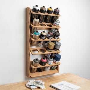 May include: A wooden spray paint can organizer mounted on a white wall. The organizer holds multiple cans of spray paint in various colors, including black, brown, yellow, and blue. The organizer is made of light-colored wood and has several shelves with circular cutouts.