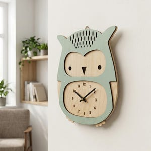 May include: A wooden owl-shaped wall clock with a light green border and a natural wood face. The clock features black hands and numbers, and is hanging on a white wall. The clock measures approximately 25 cm in height.