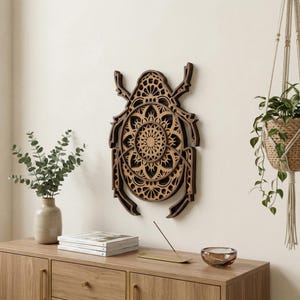 Puede incluir: Una pieza de arte mural de madera en capas con forma de escarabajo, con intrincados diseños estilo mandala. La obra de arte es principalmente marrón y negra, con recortes detallados que crean un efecto tridimensional. Está montada en una pared de color neutro.