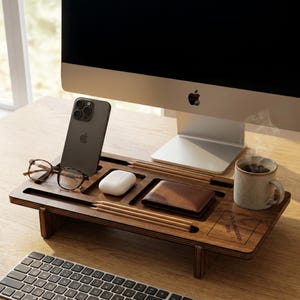 May include: A wooden desk organiser holding a phone, glasses, wallet, pencils, and a coffee mug. The organiser is positioned under a computer monitor. The phone is black, and the wallet is brown. The organiser has the text "Personalization Area".