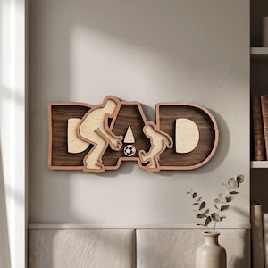 Puede incluir: Arte de pared de madera con la palabra "DAD" con temática de fútbol. Las letras son marrones con un interior de madera clara. La "A" presenta un balón de fútbol y siluetas de una persona y un niño jugando al fútbol.