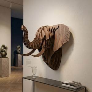Può includere: Una scultura a forma di testa di elefante in legno, montata su una parete. La scultura è realizzata con strati di legno, creando un effetto tridimensionale. L'elefante ha grandi zanne e una proboscide dettagliata. Il legno è di colore marrone caldo naturale.