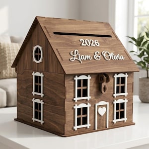 May include: A wooden house-shaped box with a slot on the roof. The box is brown with white window frames and a heart-shaped door. The text "2026 Liam & Olivia" is displayed on the roof.