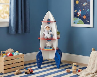 May include: A white rocket-shaped bookshelf with red shelves and blue accents. The bookshelf holds a stuffed astronaut, a star, books, and a small plant. A framed space-themed picture hangs on the blue wall. A wooden crate with toys sits on a striped rug.