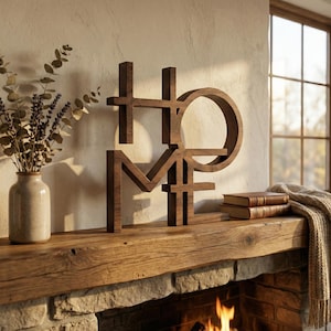 May include: A wooden "HOME" sign in a warm brown hue sits on a rustic wooden mantel. The letters are stylized and connected. A vase with dried flowers and two books are also on the mantel. A cozy blanket and a window are in the background.