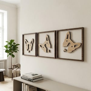 May include: Three framed wooden wall art pieces featuring dog head silhouettes. Each piece has a dark brown frame and a light wood dog design. The art is mounted on a white wall, creating a modern and minimalist aesthetic.