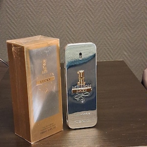 Puede incluir: Un frasco de perfume plateado y una caja dorada. El frasco tiene la inscripción "Million Lucky", y la caja "Lucky". La caja también tiene el texto "100ml 3.4FL OZ".