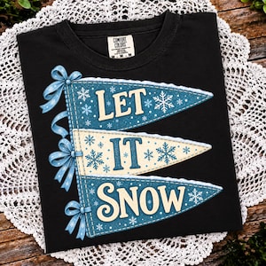 May include: A black t-shirt featuring a graphic of three stacked triangular pennants in blue and cream colors. The pennants are decorated with white snowflake patterns and light blue bows. The text "LET IT SNOW" is printed across the pennants.