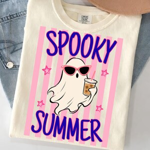 May include: A cream-colored t-shirt featuring a graphic of a white ghost wearing pink cat-eye sunglasses and holding an iced coffee. The text reads SPOOKY SUMMER in blue letters over a background of vertical pink stripes and small pink stars.