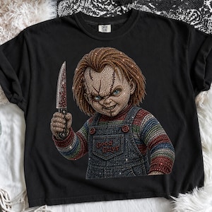 May include: A black t-shirt featuring a rhinestone-embellished graphic of a doll with red hair, blue eyes, and a scarred face. The doll wears blue denim-style overalls with the text Good Guys and holds a silver knife with red accents.