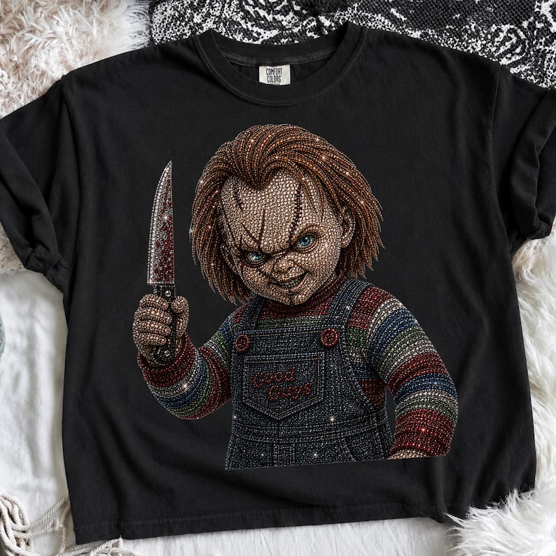 May include: A black t-shirt featuring a rhinestone-embellished graphic of a doll with red hair, blue eyes, and a scarred face. The doll wears blue denim-style overalls with the text Good Guys and holds a silver knife with red accents.