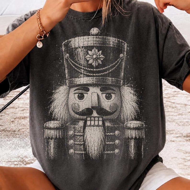 May include: A charcoal gray short-sleeve t-shirt featuring a large, distressed white graphic print of a nutcracker soldier. The design displays the nutcracker from the chest up, showcasing a tall hat, a mustache, and a long, textured beard.