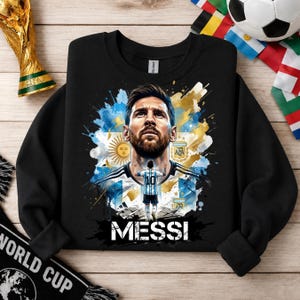 May include: A black long-sleeve sweatshirt featuring a graphic print of Lionel Messi. The design includes a portrait of Messi, a smaller figure of him in an Argentina jersey with the number 10, the Argentina flag, and the text MESSI in white.
