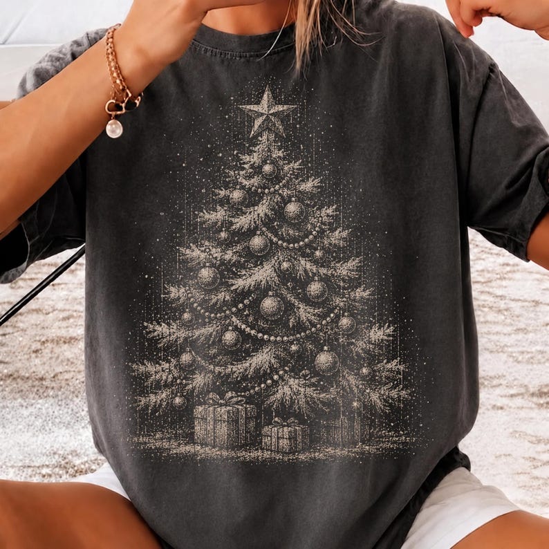 May include: A charcoal gray short-sleeve t-shirt featuring a detailed, light-colored graphic print of a decorated Christmas tree. The tree is adorned with ornaments, garlands, and a star topper, with several wrapped gift boxes placed at the base.