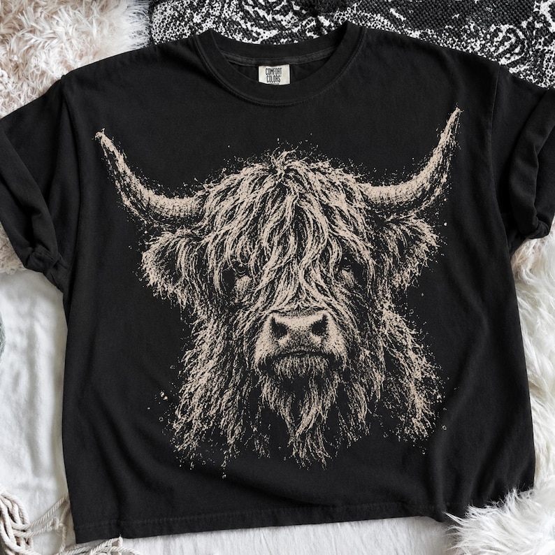 May include: A black short-sleeve t-shirt featuring a large, detailed beige graphic of a Highland cow head with prominent horns. The shirt has a crew neckline and a Comfort Colors brand label printed on the inside of the collar area.