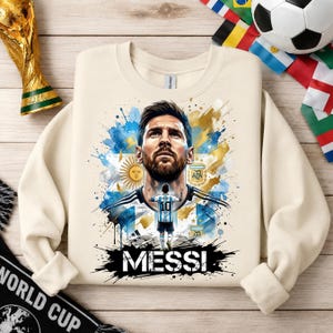 May include: A cream-colored crewneck sweatshirt featuring a graphic print of Lionel Messi. The design includes a portrait of Messi, a smaller figure wearing a number 10 jersey, the AFA logo, a sun emblem, and the text MESSI in bold black letters.