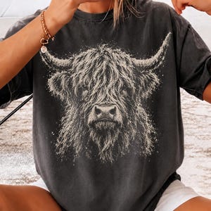 May include: A charcoal grey short-sleeve t-shirt featuring a large, detailed graphic print of a Highland cow head in a light beige color. The cow has long, shaggy fur and prominent curved horns, centered on the front of the garment.