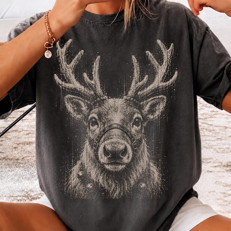 May include: A charcoal gray short-sleeve t-shirt featuring a detailed, light-colored graphic print of a reindeer head with large antlers. The reindeer is depicted with a harness around its snout and small bells around its neck in a sketch style.