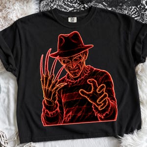 May include: A black t-shirt featuring a neon red graphic illustration of a character wearing a fedora hat and a striped sweater, with a clawed glove on one hand. The design has a glowing outline effect against the dark fabric of the shirt.